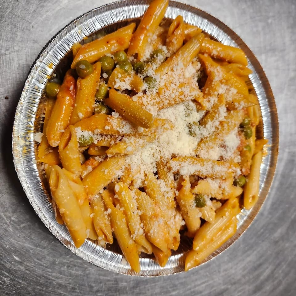 Penne pasta with tomato sauce, green peas, and grated cheese in a foil takeout container.