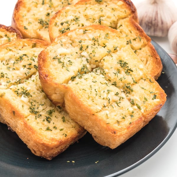 Golden-brown garlic bread slices topped with herbs on a dark plate, with garlic bulbs in the background.