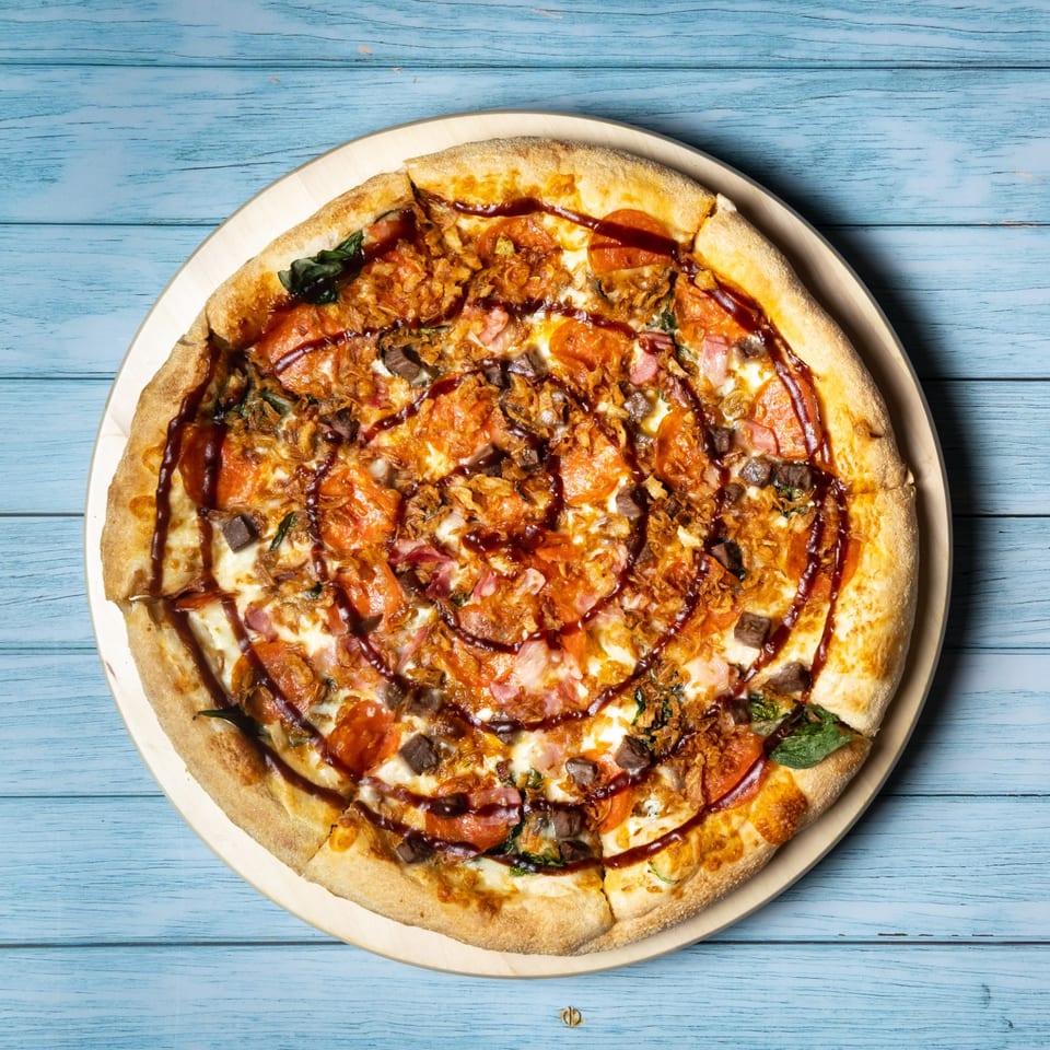 Tommy's Pizza with pepperoni, ham, beef, crispy onions, and BBQ sauce drizzle on a blue wooden table.